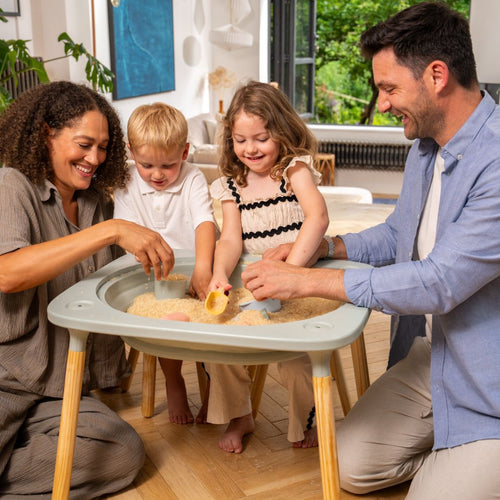 Two adults and two children play with sand and toy scoops at the TP Toys Active-Tots Pikler Style 4 in 1 Wooden Play Table (FSC® certified) in a bright, modern living room.