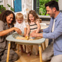 Two adults and two children play with sand and toy scoops at the TP Toys Active-Tots Pikler Style 4 in 1 Wooden Play Table (FSC® certified) in a bright, modern living room.