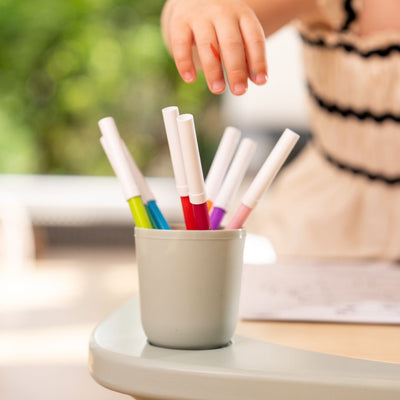 A child's hand reaches for markers in a cup on the TP Active-Tots Pikler Style 4 in 1 Wooden Play Table by TP Toys, FSC® certified, with greenery softly blurred in the background.