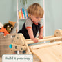 A young child climbs on the TP Toys Active-Tots Pikler Style Wooden Climbing Triangle & Slide (FSC® certified) in a playroom with toys, books, and a light blue wall. A sign in the corner reads: Build it your way.