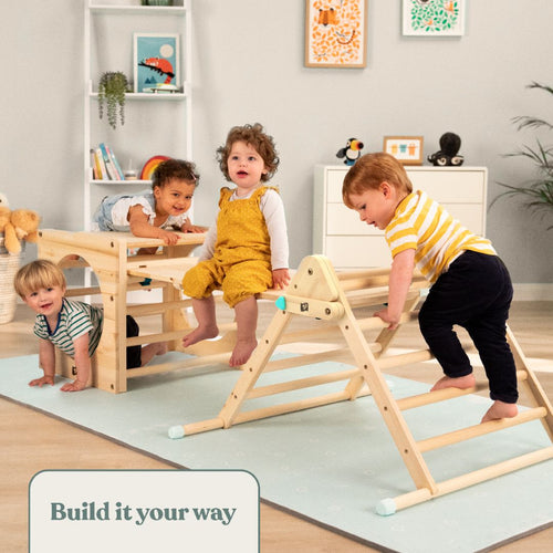 Three young children play on the TP Active-Tots Pikler Style Wooden Climbing Triangle by TP Toys in a cozy living room with bookshelves, toys, and animal wall art. A sign reads, "Build it your way." The climbing triangle is FSC® certified.