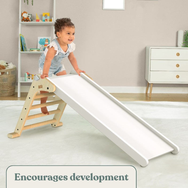 A toddler climbs the TP Toys Active-Tots Pikler Style Folding Wooden Slide—FSC® certified—in a playroom, smiling with toys and shelves behind. Caption: Encourages development and indoor play.