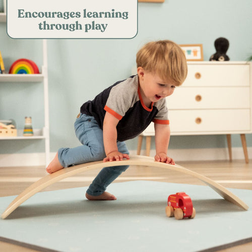 A young child balances on the TP Toys TP Active-Tots Pikler Style Wooden Balance Board - FSC® certified while reaching for a red toy train in a playroom, beneath a sign that reads: Encourages learning through play.