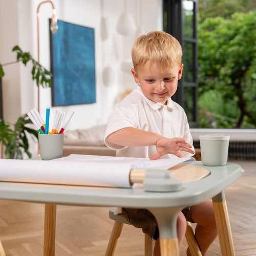 A young boy sits at a TP Active-Tots Play Table Paper Roll Accessory, drawing on a paper roll with a cup of markers nearby.