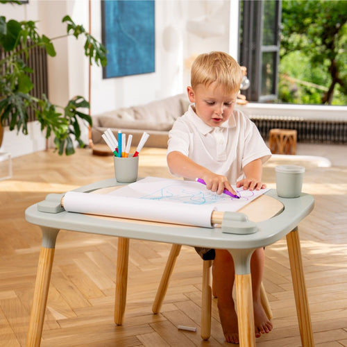 A young boy sits at a table drawing on a TP Active-Tots Play Table Paper Roll Accessory attached to the tabletop.