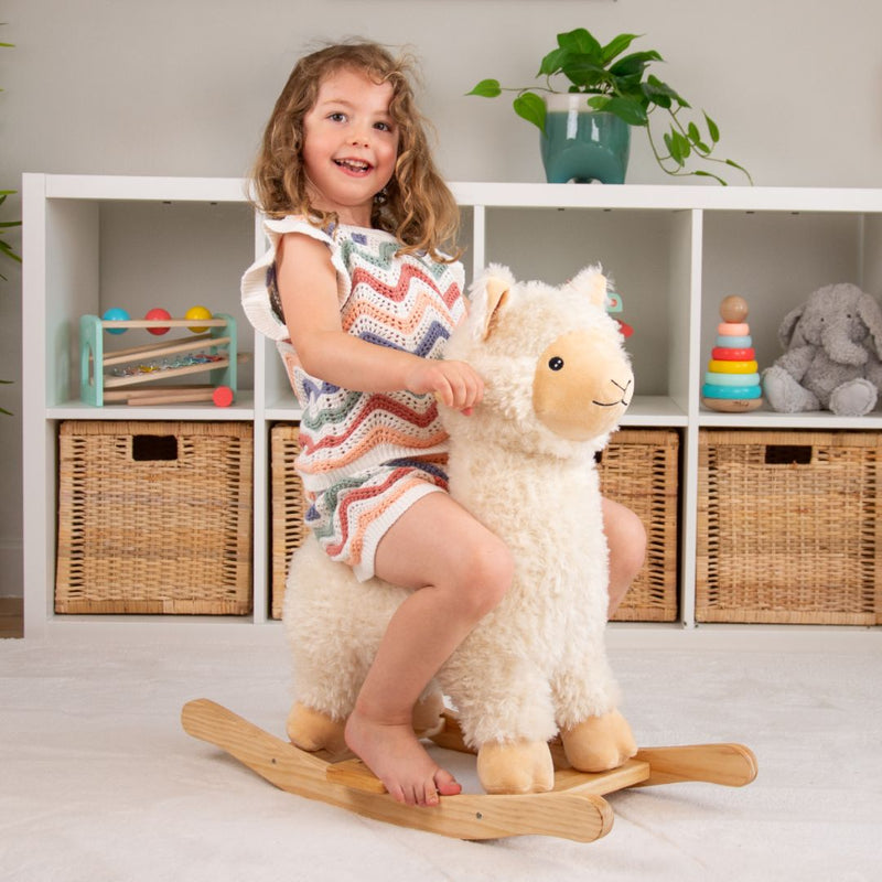 A smiling child rides the TP Toys Active-Tots Rocking Llama, a soft plush toddler rocker, in a playroom decorated with shelves, baskets, and colorful toys in the background.