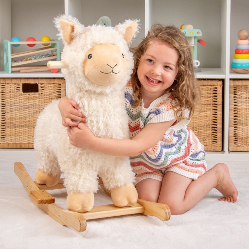 A young child smiles and hugs the TP Toys Active-Tots Rocking Llama in a playroom, with wicker baskets and colorful toys neatly arranged on shelves in the background.