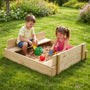 Two young children play with the TP Wooden Lidded Sandpit by TP Toys, FSC® certified, on a grassy lawn surrounded by greenery and flowers.
