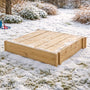The TP Toys TP Wooden Lidded Sandpit - FSC® certified sits closed on a snow-dusted lawn, waiting for warmer days to invite outdoor play back to the garden.