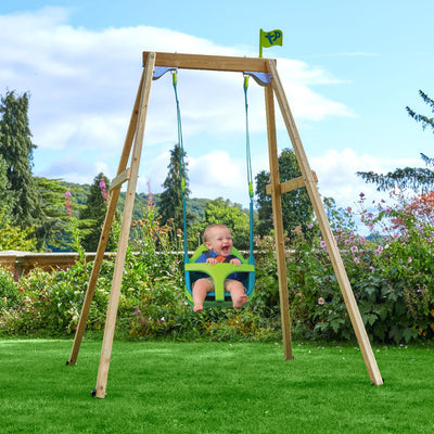 A baby sits in the TP Toys TP Forest Wooden Single Swing with Quadpod (FSC® certified) on a grassy lawn, surrounded by shrubs, trees, and a blue sky.