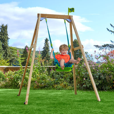 A young child smiles while swinging on the TP Toys TP Forest Wooden Single Swing with Quadpod, made from FSC® certified wood, in a grassy garden surrounded by colorful flowers and trees.