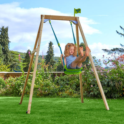 A young girl with curly hair swings on the TP Toys TP Forest Wooden Single Swing with Quadpod—FSC® certified—in a garden surrounded by grass, flowers, and trees under a partly cloudy sky.