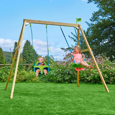Two children swing on the TP Toys TP Forest Wooden Double Swing with Quadpod, made from FSC® certified wood, in a grassy garden filled with flowers and trees under a blue sky.