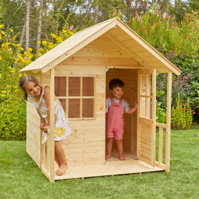 Two young children play in the TP Toys TP Forest Cabin Wooden Playhouse, made from FSC® certified wood, on a grassy lawn surrounded by green plants and yellow flowers.