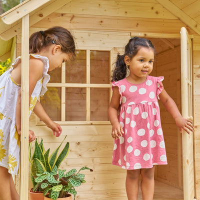 Two young girls play by the TP Toys TP Forest Cabin Wooden Playhouse – FSC® certified; one enters while the other stands at the doorway. A potted plant sits beside this charming garden cabin.