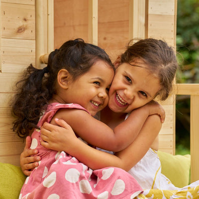 Two young girls hug and smile together in front of the TP Toys TP Forest Cabin Wooden Playhouse, made from FSC® certified wood.
