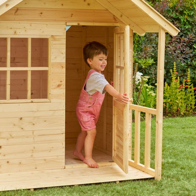 A young child smiles as they stand in the doorway of the TP Toys TP Forest Cabin Wooden Playhouse – FSC® certified, holding open the door to their cozy retreat on a grassy lawn.