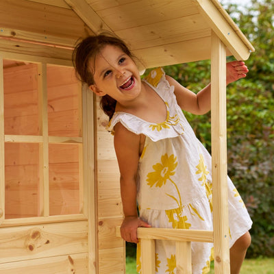 A young child smiles while standing in the doorway of the TP Forest Cabin Wooden Playhouse by TP Toys, crafted from FSC® certified wood and set outdoors.