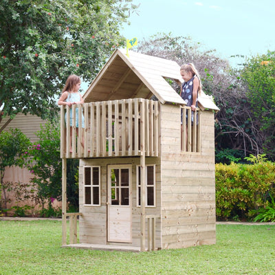 Two young girls stand on the balcony of a TP Toys TP Loft Two Storey Wooden Playhouse - FSC® certified, set in a grassy backyard surrounded by trees and bushes.