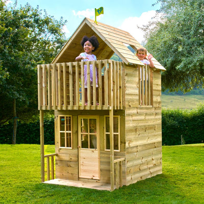 Two children smile on the upper balcony of the TP Loft Two Storey Wooden Playhouse by TP Toys, set in a grassy yard with trees behind. This playhouse is FSC® certified.