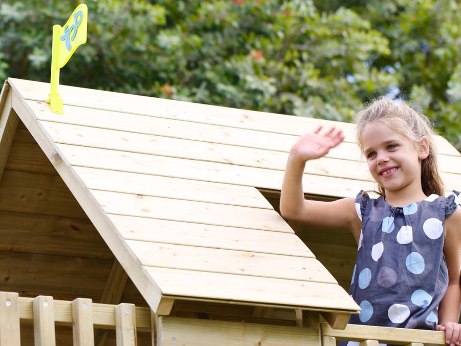 A young girl in a polka dot dress smiles and waves on the porch of the TP Toys TP Loft Two Storey Wooden Playhouse (FSC® certified), set outdoors with trees in the background.