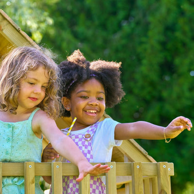 Two young girls smile and reach over the railing of the TP Toys TP Loft Two Storey Wooden Playhouse - FSC® certified, standing outdoors with green foliage in the background.