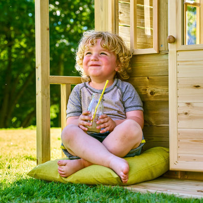 A young child with curly blond hair sits cross-legged on a cushion outside the TP Toys TP Loft Two Storey Wooden Playhouse – FSC® certified, sipping from a straw bottle and enjoying the sunshine.