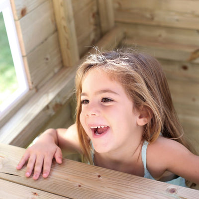 A young child with long light brown hair smiles and looks out the window of the TP Toys Loft Two Storey Wooden Playhouse, FSC® certified, on a sunny day.