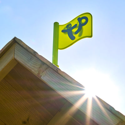 A bright green flag with a blue tp logo flies atop the TP Toys TP Loft Two Storey Wooden Playhouse - FSC® certified, with sunlight shining in the background.