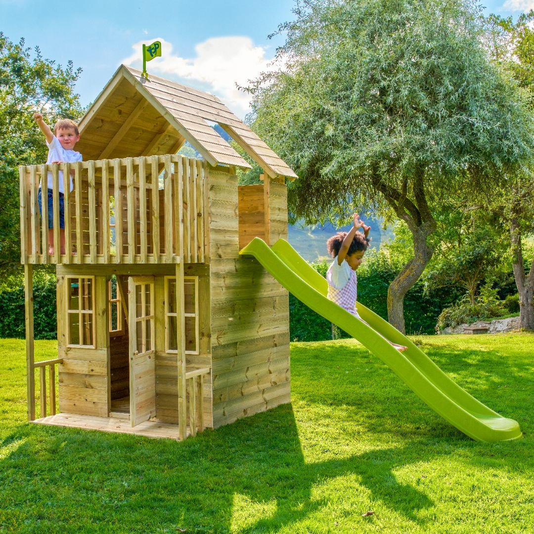 Two children enjoy the TP Toys TP Skye Two Storey Wooden Playhouse (FSC® certified); one stands on the balcony while the other slides down the green wavy slide, all set on a grassy lawn with trees in the background.