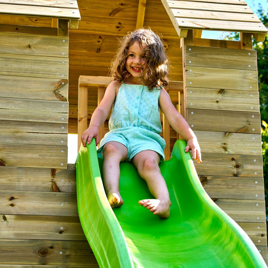 A young child with curly hair in a light blue outfit slides down a green wavy slide attached to the TP Skye Two Storey Wooden Playhouse—FSC® certified by TP Toys, enjoying outdoor fun.