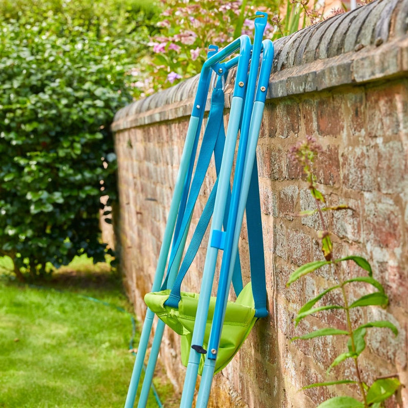 The TP Toys TP Foldaway Baby Swing is folded and leaning against a brick wall in a grassy outdoor area with nearby plants.