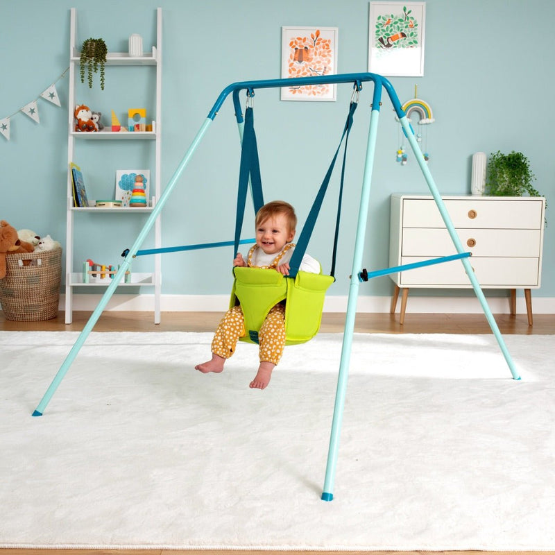 A smiling baby enjoys the TP Toys TP Foldaway Baby Swing in green, suspended from a blue frame in a colorful nursery filled with toys and artwork.