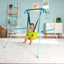 A smiling baby enjoys the TP Toys TP Foldaway Baby Swing in green, suspended from a blue frame in a colorful nursery filled with toys and artwork.