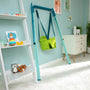 The TP Toys TP Foldaway Baby Swing, in green, hangs from a blue swing frame in a light blue nursery with a white shelf, dresser, and toys.