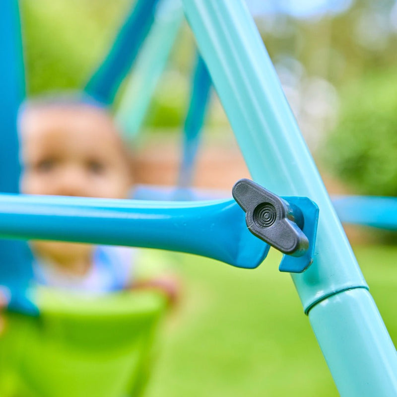 Close-up of the TP Toys TP Foldaway Baby Swings blue metal frame with a black knob; a young child sits in the green seat, blurred in the outdoor background.