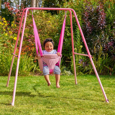 A young child smiles in a TP Toys TP Pink Foldaway Baby Swing, wearing a headband and patterned pants, surrounded by green grass and plants in a garden.