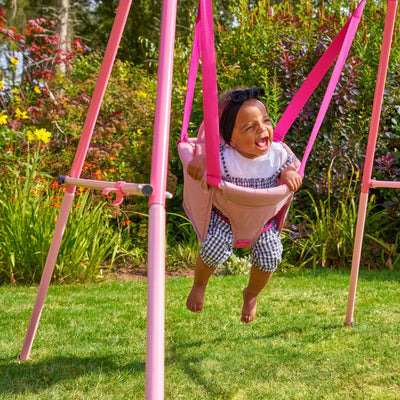 A young child smiles while swinging in the TP Toys TP Pink Foldaway Baby Swing outdoors, surrounded by green grass and colorful flowers.