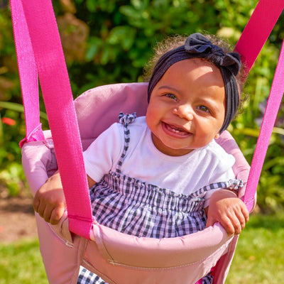 A smiling baby in a black headband and checkered dress sits in the TP Toys TP Pink Foldaway Baby Swing, surrounded by lush greenery outdoors.