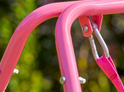 Close-up of the TP Pink Foldaway Baby Swing by TP Toys, featuring a pink metal frame with a carabiner attaching a pink strap, set against a blurred green and purple background.