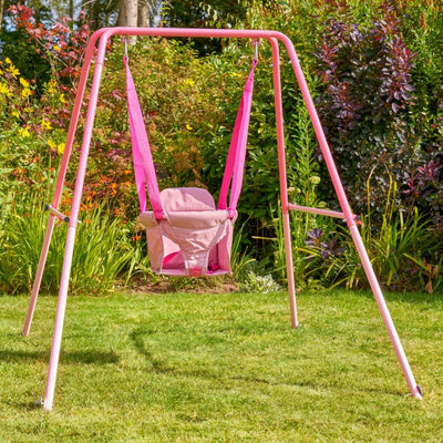The TP Toys TP Pink Foldaway Baby Swing features a fabric seat and is perfect for indoor or outdoor use, shown here set on a grassy lawn surrounded by green plants in a garden.