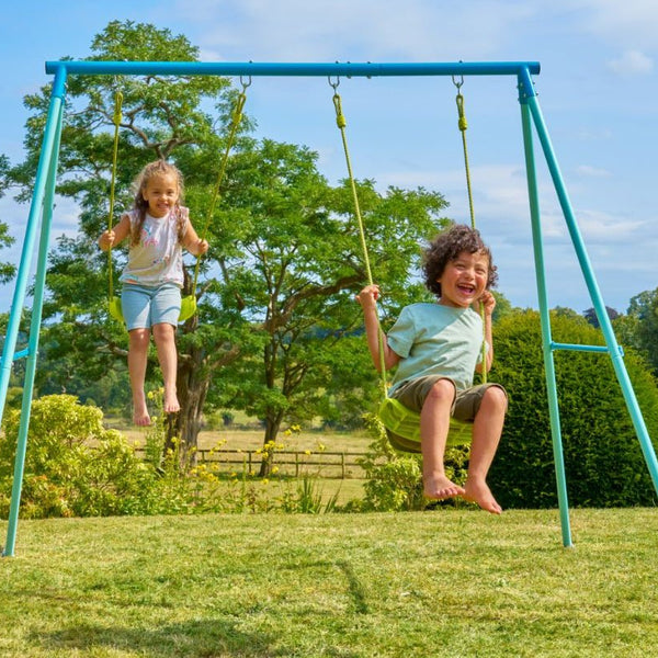 Two children are playing on a TP Toys TP Double Metal Swing Set with injection-moulded seats in a grassy backyard surrounded by trees and greenery.