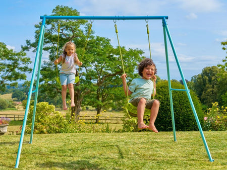 Two children are playing on a TP Toys TP Double Metal Swing Set with injection-moulded seats in a grassy backyard surrounded by trees and greenery.