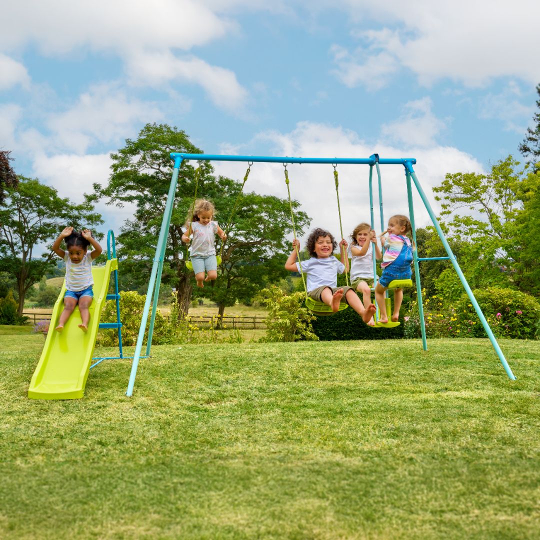 Five kids play outside on the TP Toys TP Metal Double Swing Slide & Glide set: two use the adjustable swing seats, while three enjoy the slide. The playground is set on grass with trees and a blue sky in the background.