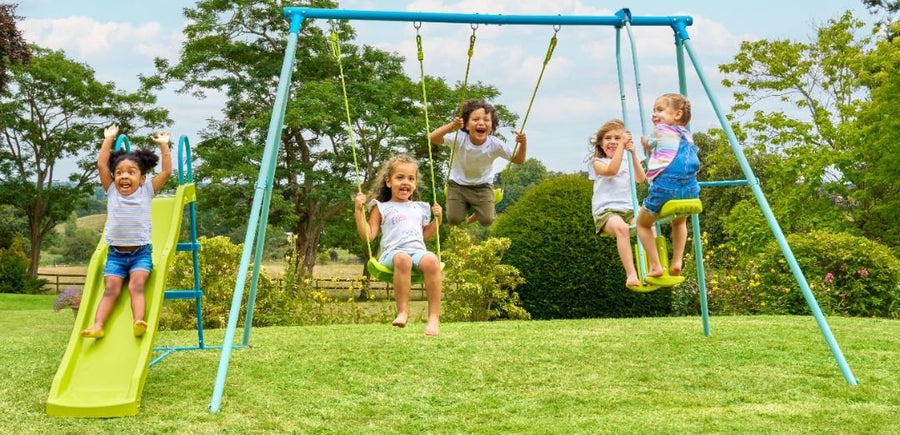 Five children enjoy the TP Toys TP Metal Double Swing Slide & Glide set in a grassy yard: two on adjustable swings, two on the slide, and one standing. Trees and clouds fill the background.
