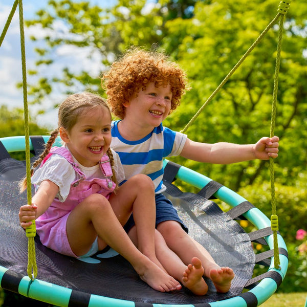 Two young children smile and enjoy a sunny day together on the TP Toys Giant Nest Metal Swing Set in a green garden.
