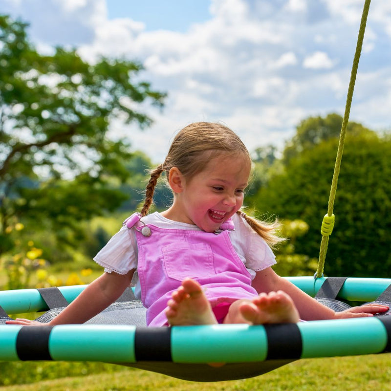 A young girl with pigtails, in a pink overall, smiles as she sits on the TP Toys TP Giant Nest Metal Swing Set in a sunny green garden.