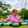 A young girl with pigtails, in a pink overall, smiles as she sits on the TP Toys TP Giant Nest Metal Swing Set in a sunny green garden.