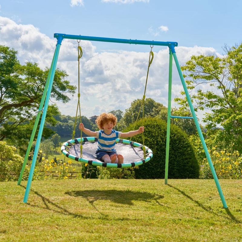 A young child enjoys the TP Toys TP Giant Nest Metal Swing Set in a grassy outdoor area, with trees and a cloudy sky in the background.