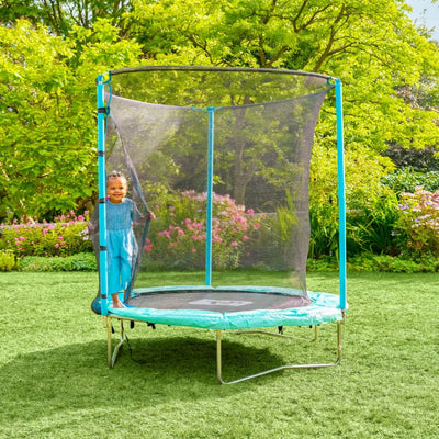A young child smiles inside a TP Toys TP UP 6ft Trampoline on a grassy lawn, enjoying its safety features with trees and flowers in the background.